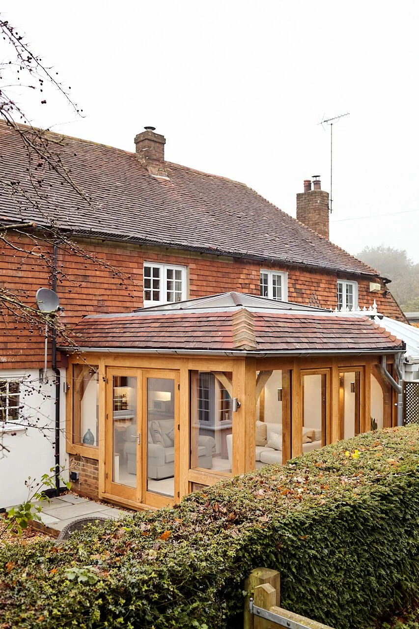 The image shows a house with a brick exterior and a tiled roof, featuring an oak framed orangery extension, with large glass doors framing a cozy indoor seating area.