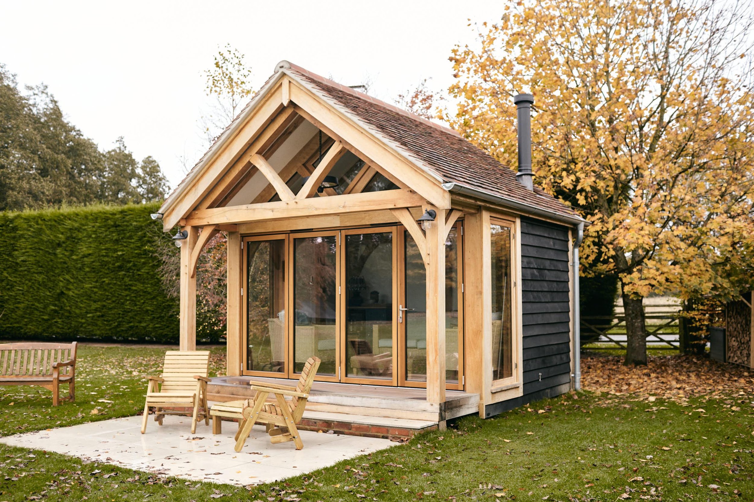 An oak framed garden room cabin studio with black featheredge cladding, and large glass sliding doors and an outdoor patio with chairs, surrounded by trees with fall foliage.