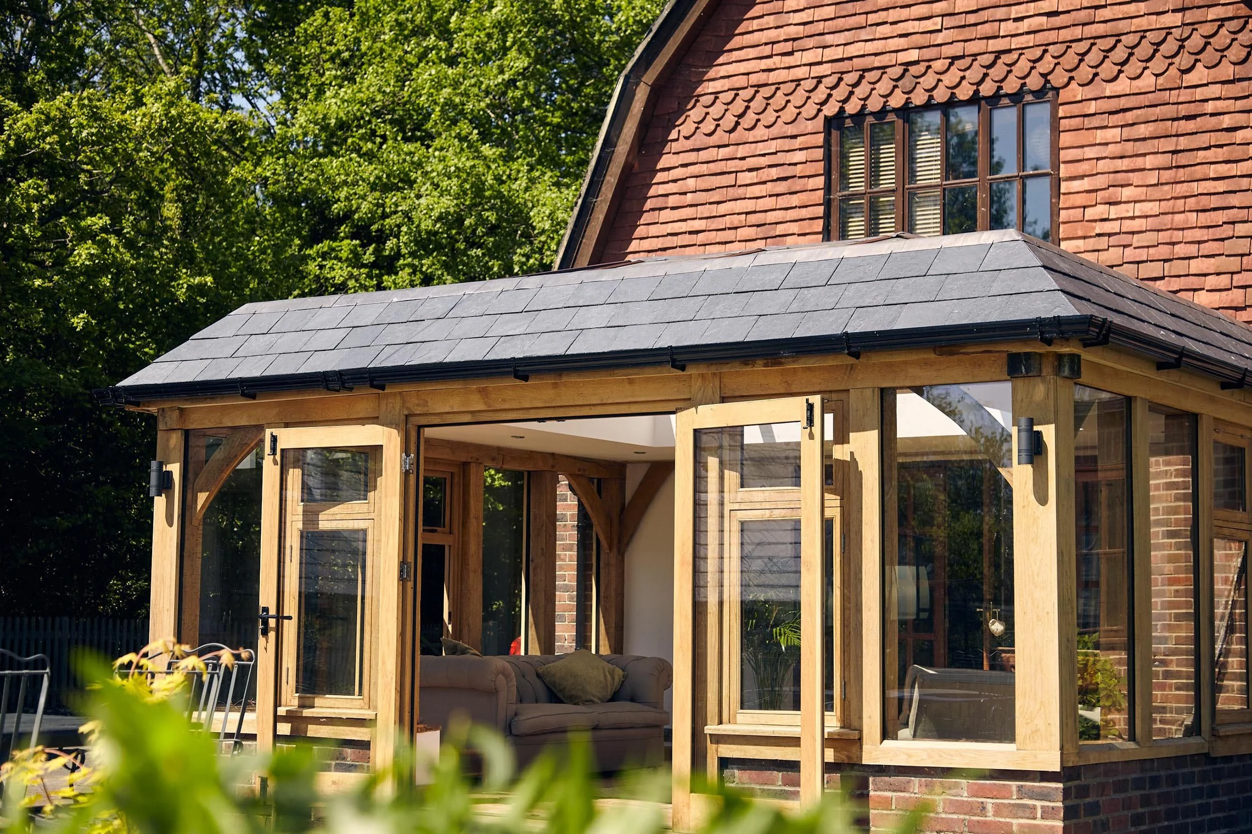 A house with a brick exterior and an oak framed orangery extension with mansard style roof, surrounded by green trees, on a sunny day.