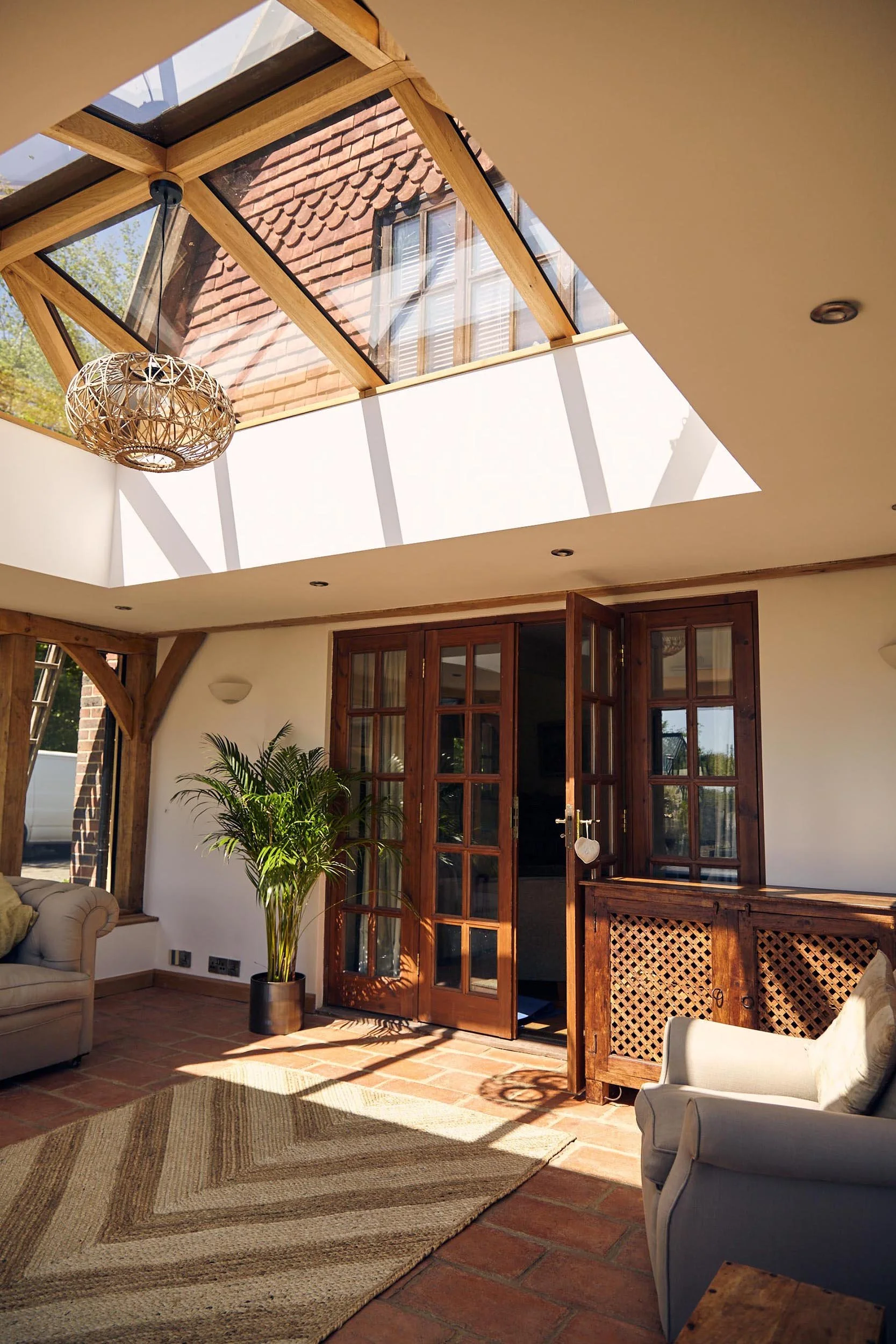 Living room in an oak framed orangery with an oak framed skylight roof lantern, glass doors, a potted plant, sofas, a patterned rug, and sunlight streaming in.