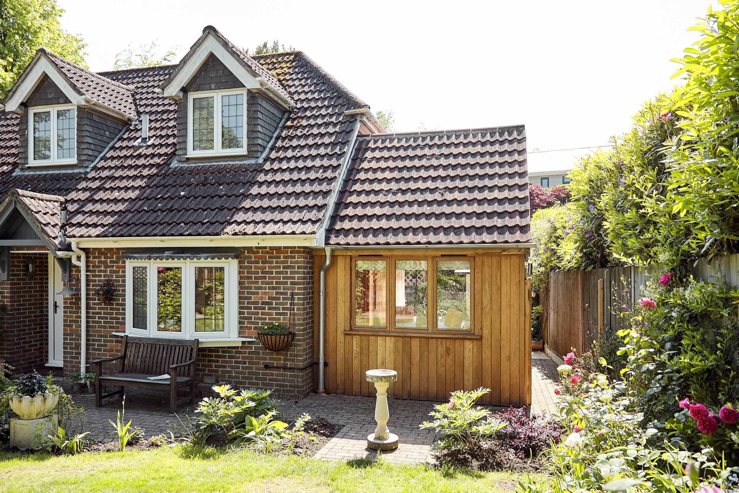 Front view of a brick house with a tiled roof and a small oak framed extension with vertical oak channel cladding, surrounded by a garden with plants and flowers.
