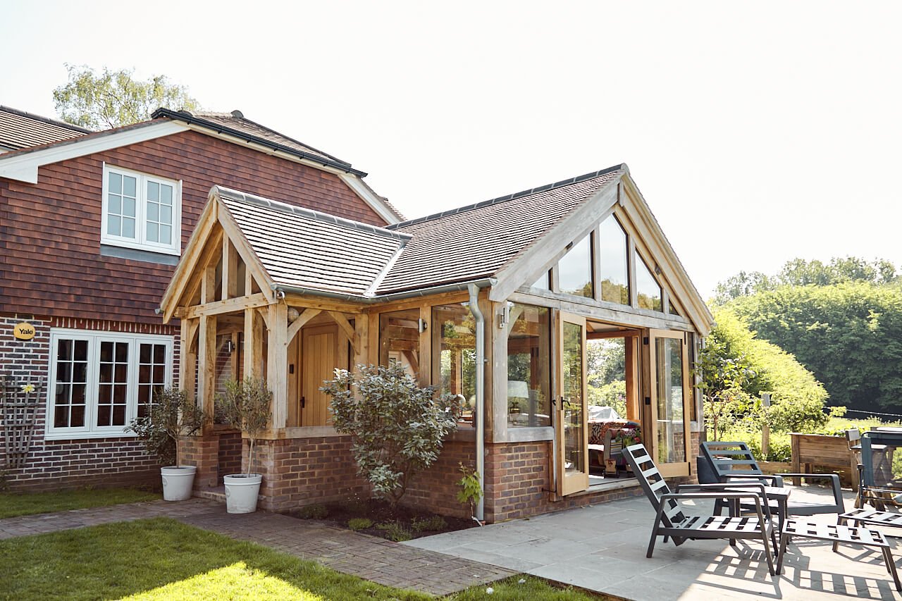 Exterior view of a house with a green oak framed extension, featuring large glass windows and patio chairs outside in a garden setting.