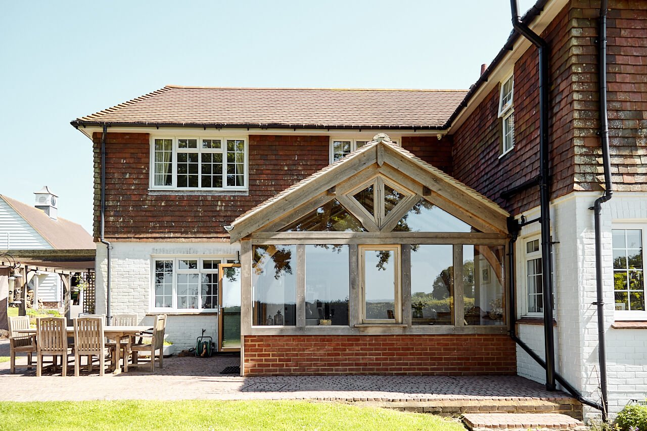 Back view of a brick house with an oak framed extension sunroom in the garden area, outdoor patio furniture, and neighboring houses in the background.