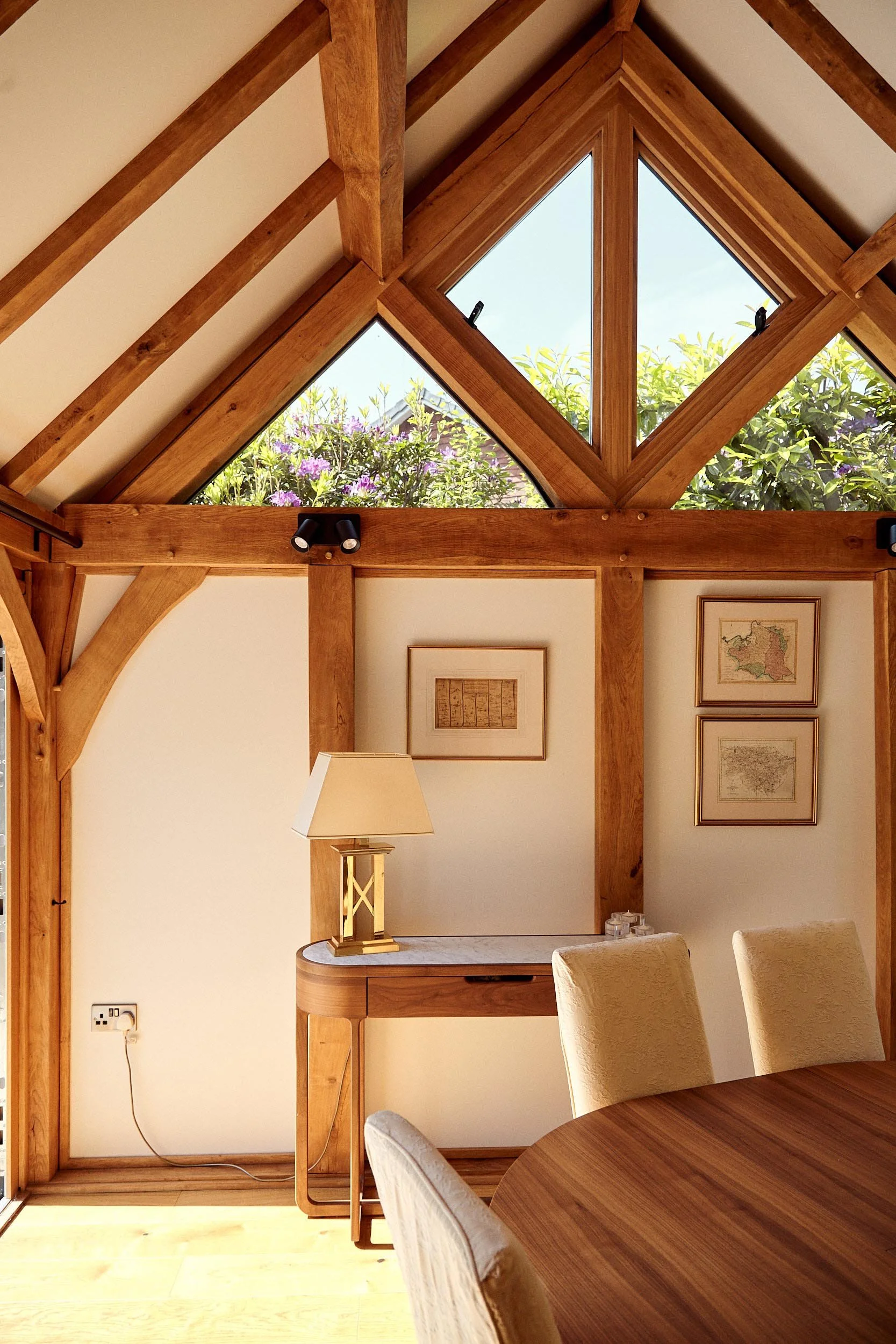 A cozy dining area in an oak framed house extension, featuring a wooden table and chairs, a side table with a lamp, framed art on cream walls, large diamond-shaped windows with a view of greenery and purple flowers outside, and exposed wooden beams o