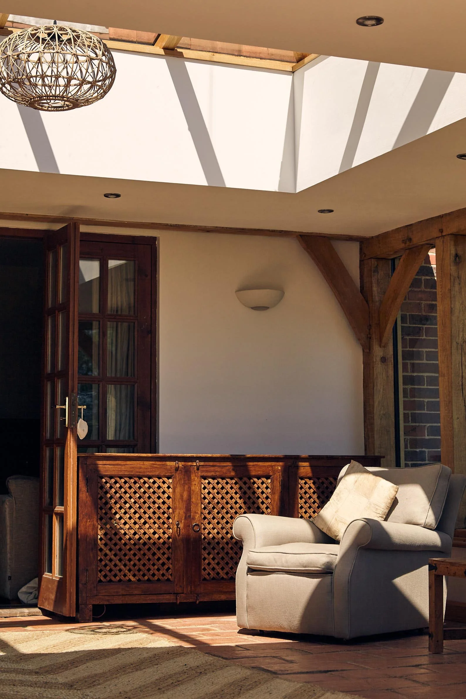Sunlight coming through a glazed oak skylight ceiling illuminating a cozy indoor space in an oak framed orangery extension, with a beige armchair, decorative pillow, wooden cabinet with lattice doors, and a brick wall with wooden beams.