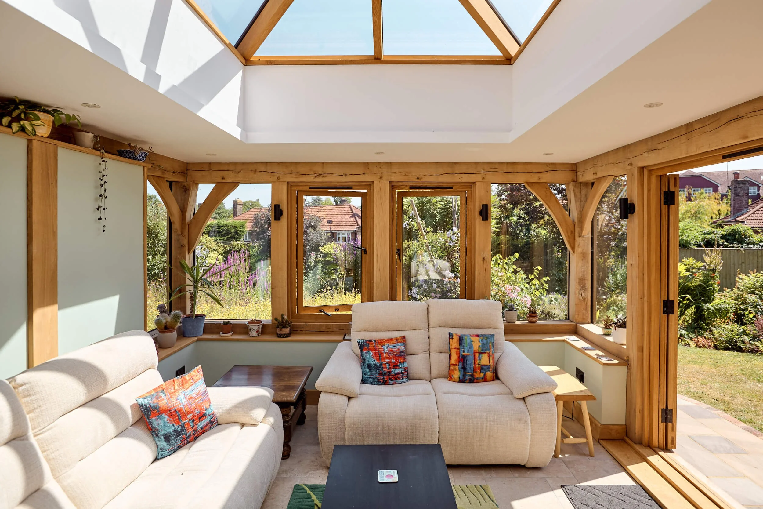 Living room with large windows, wooden framing, and a skylight ceiling. Outdoor patio with patio furniture, plants, and a garden shed visible through the windows.