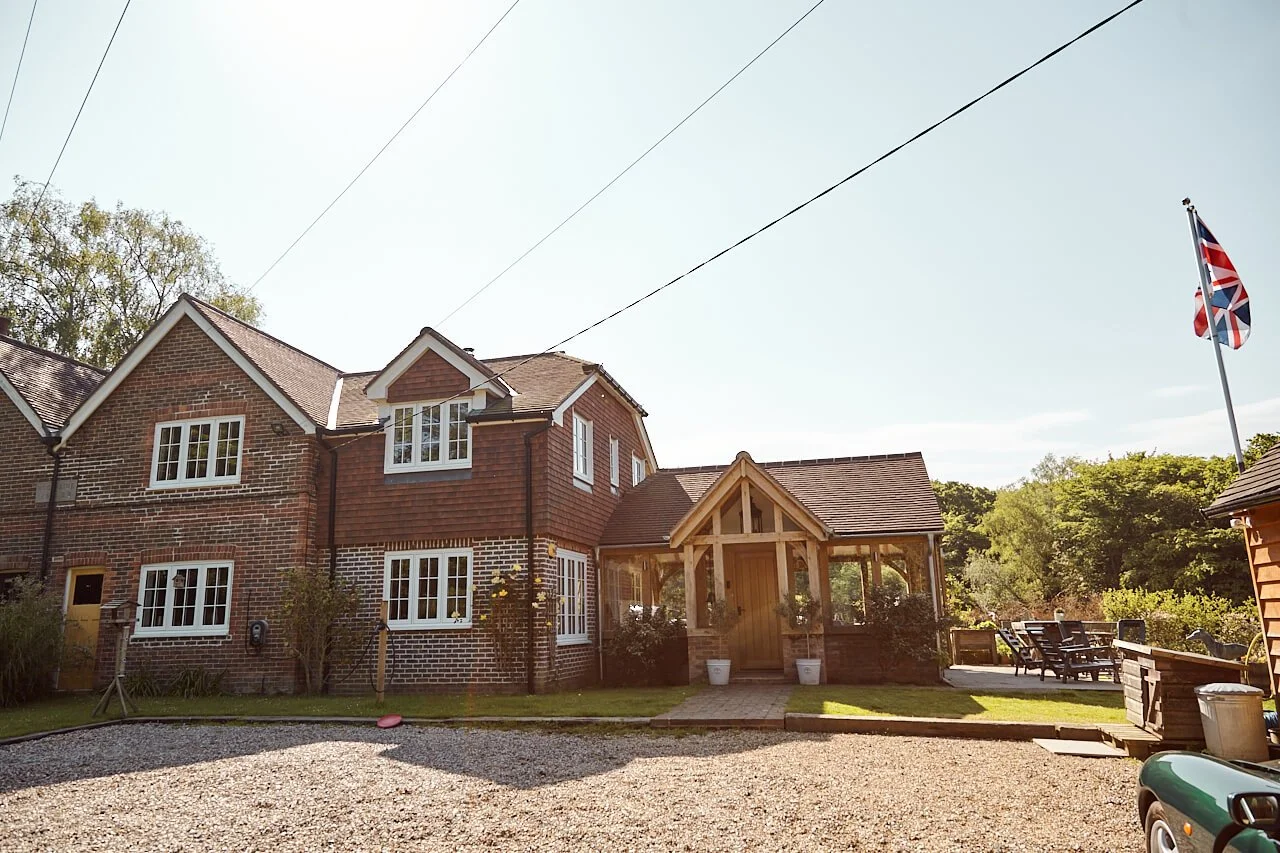 A two-story brick house with an oak framed extension and oak framed front porch, white-framed windows, and a tiled roof. There is outdoor seating in the yard, a flagpole with a Union Jack flag, and trees in the background.