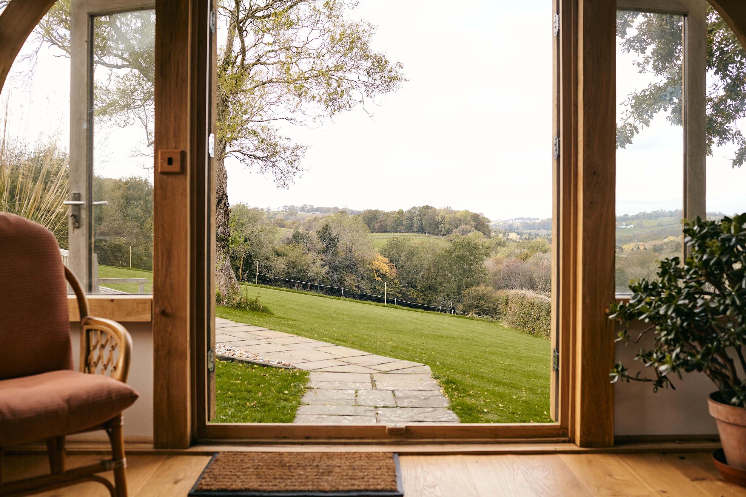 View of a lush green lawn and rolling hills seen through open wooden sliding glass doors with a plant on the right and a chair on the left inside a home.