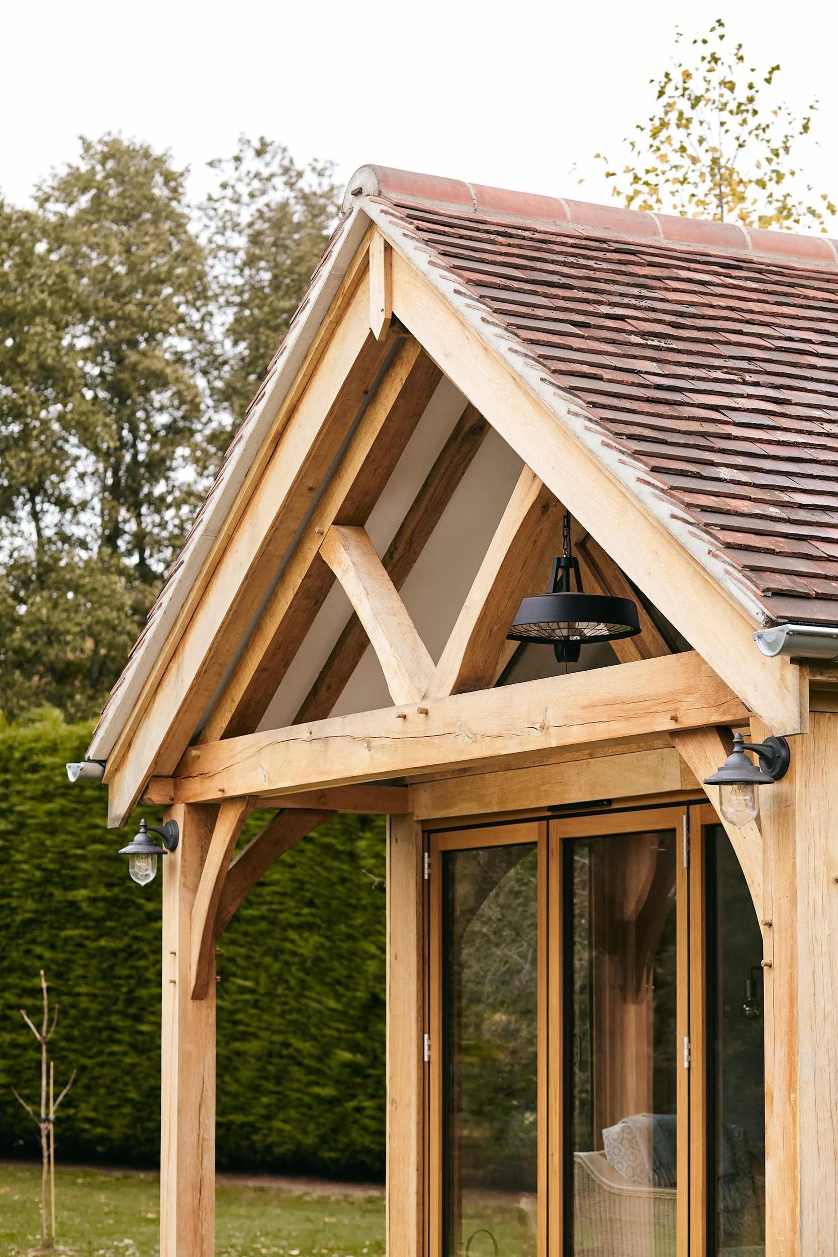 A small oak framed garden room with black painted cladding and a sloped shingle roof, situated in an outdoor area with trees and fallen leaves. The house has large glass sliding doors and a porch with outdoor chairs.