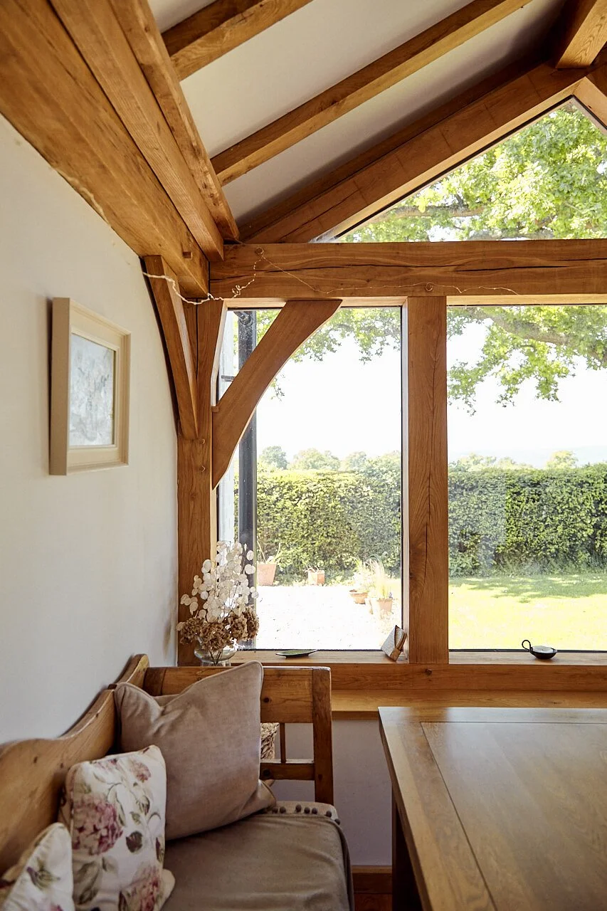 Interior of a room featuring a large window with wooden framing, a wooden ceiling with beams, and a wooden table, in an oak framed extension. There is a beige sofa with floral pillows and a vase with dried flowers on a side table. Outdoor view includ