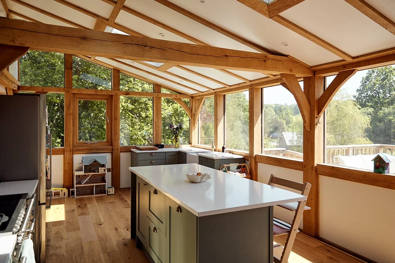Sunlit kitchen in an oak framed lean-to roof extension, with large windows, wooden beams, and a central island with a white countertop.