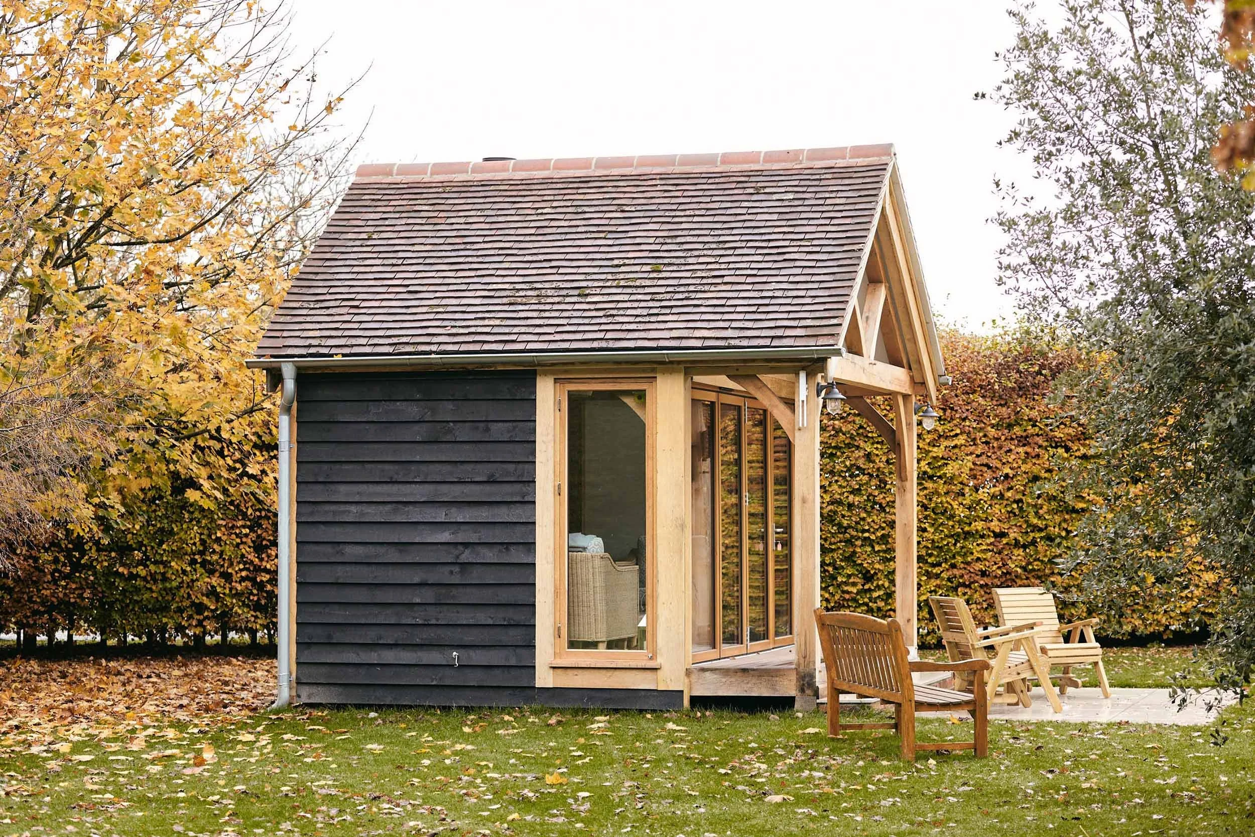 A small oak framed garden room with black painted cladding and a sloped shingle roof, situated in an outdoor area with trees and fallen leaves. The house has large glass sliding doors and a porch with outdoor chairs.