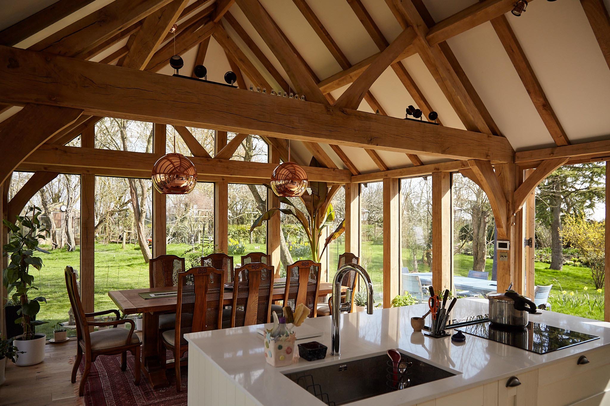 Sunlit oak framed kitchen extension with a dining table, chairs, and kitchen island, overlooking a lush back garden with trees and outdoor furniture.