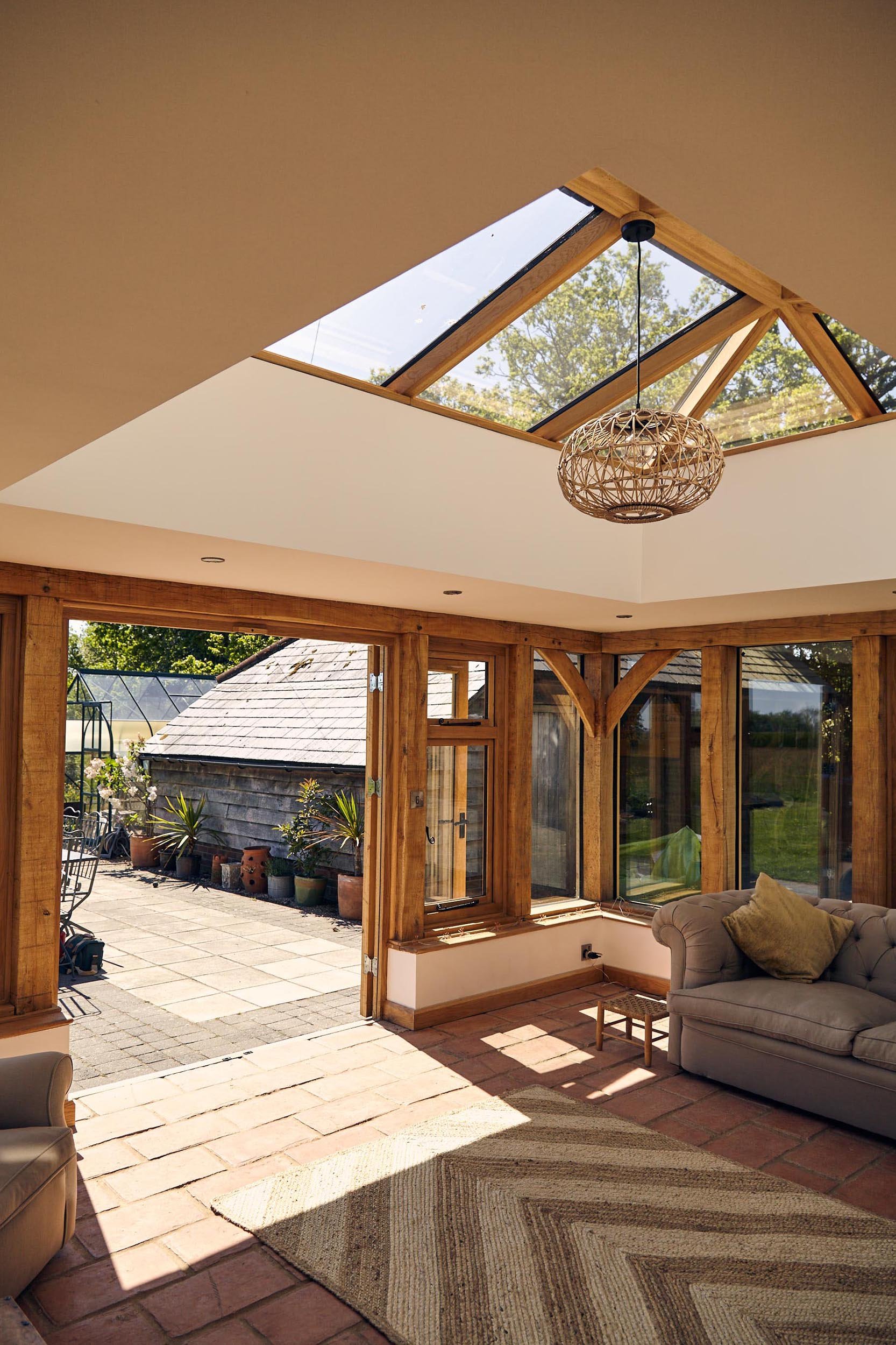 Sunroom within an oak framed orangery  extension, with a large oak skylight, wooden trim, and a view of a patio with potted plants and outdoor furniture.