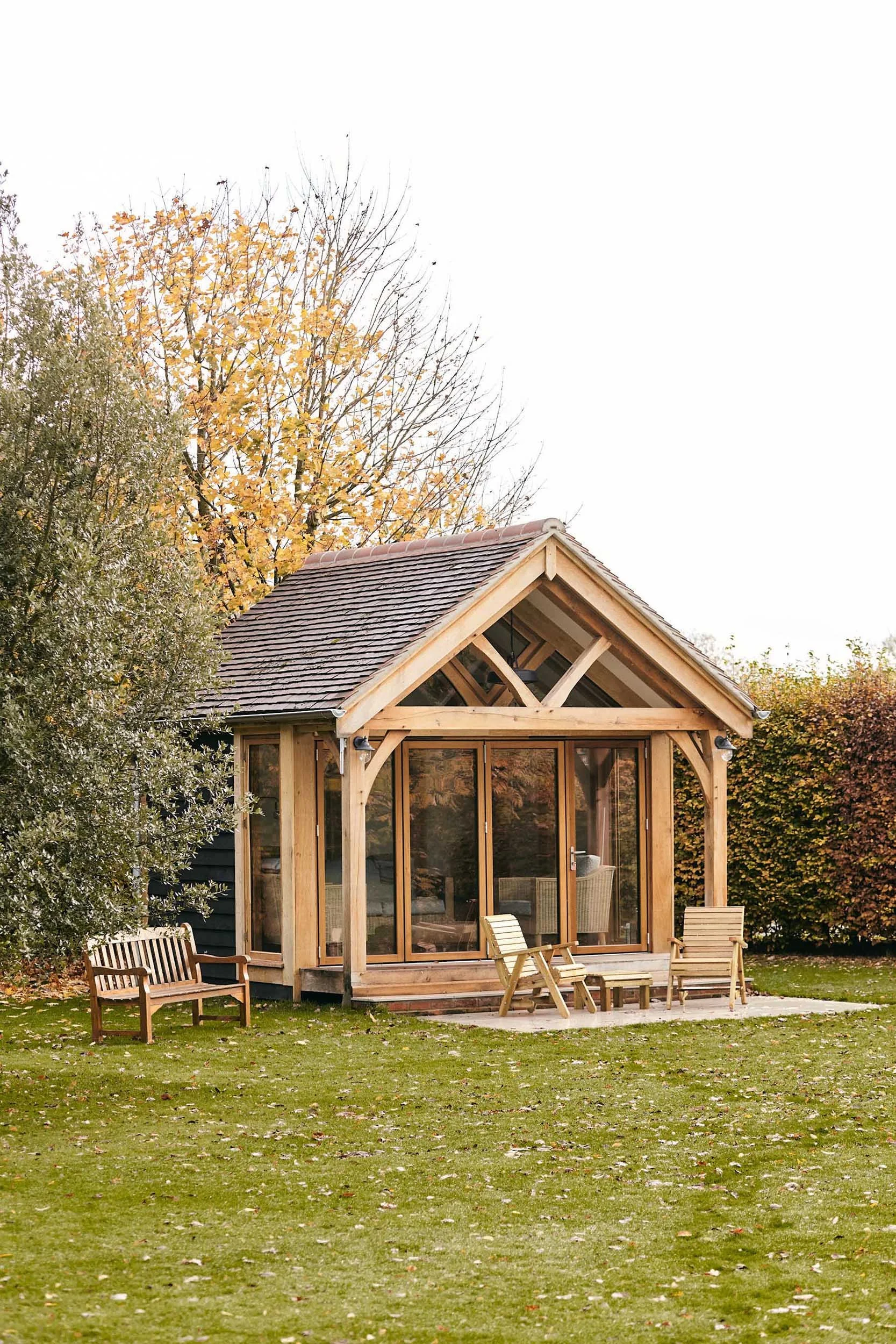 A small oak framed garden room with black painted cladding and a sloped shingle roof, situated in an outdoor area with trees and fallen leaves. The house has large glass sliding doors and a porch with outdoor chairs.