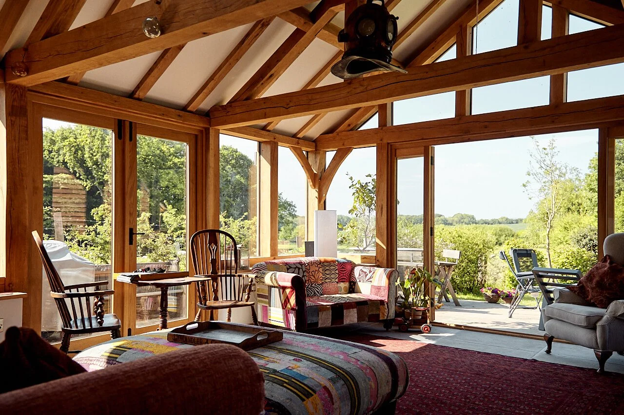 Sunlit living room in an oak framed extension with wooden beams, large windows, and a view of lush greenery outside. Furniture includes a patchwork sofa, armchair, and wooden chairs around a small table.