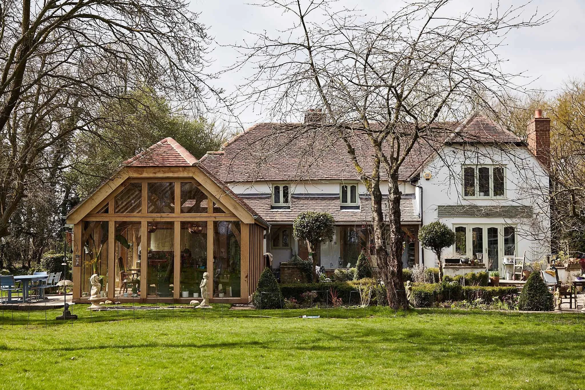A two-story house with an oak framed glazed kitchen extension on one side, surrounded by trees and a well-maintained lawn. There are small sculptures and garden furniture in the yard.