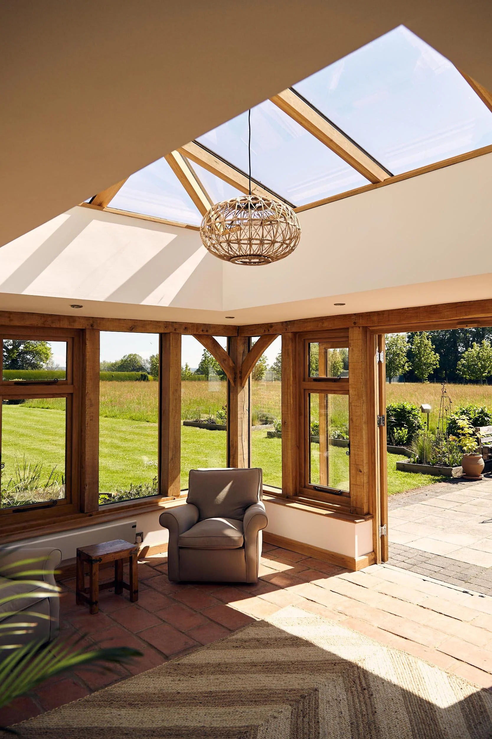A sunlit sunroom in an oak framed orangery extension with large windows showing a green field outside, a beige armchair, a small wooden side table, a woven light fixture hanging from the ceiling, and a brick floor.