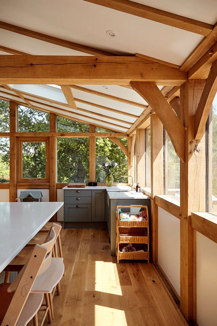A sunlit kitchen in an oak framed home extension, with wooden framing, grey cabinets, white countertop, and large windows showing greenery outside.