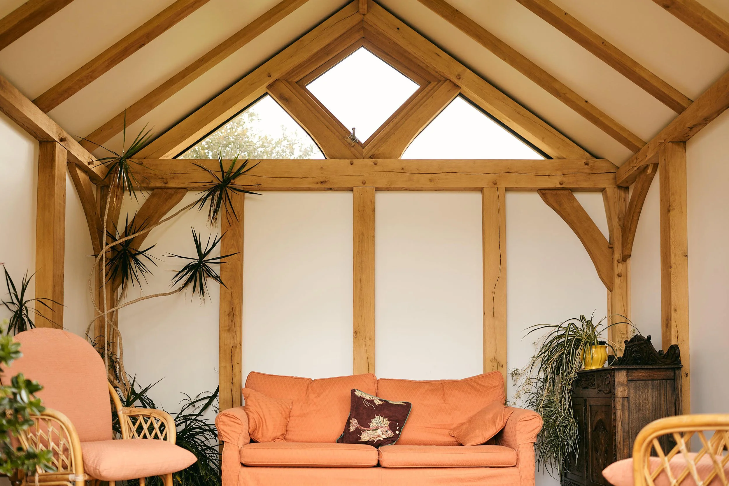 Living room in an oak framed garden room, with coral sofa, rattan chairs, wooden beams, and skylight, decorated with plants and a decorative pillow.