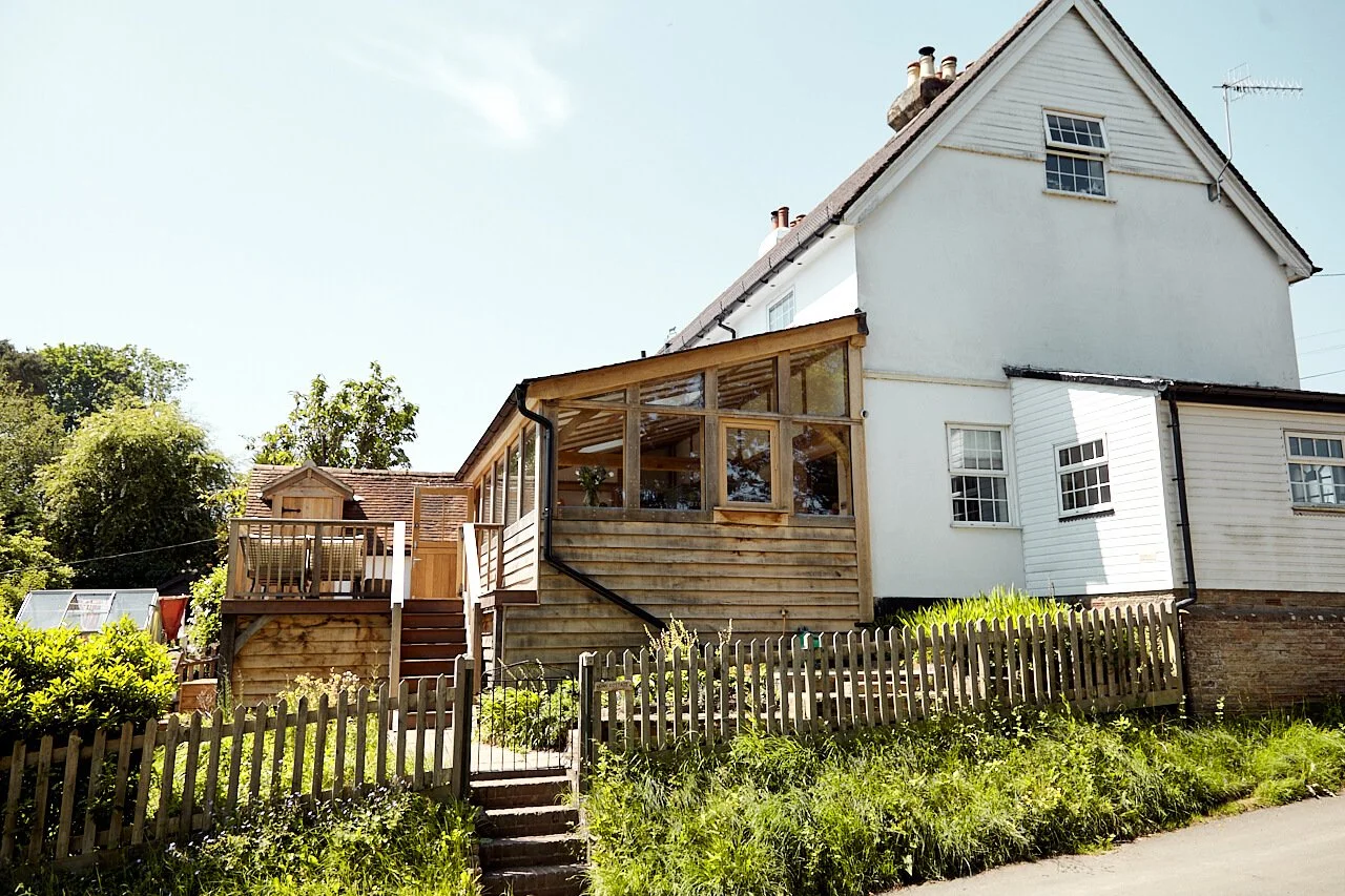 A white house with a glass-enclosed oak framed extension and a lean-to sloped roof, surrounded by a wooden fence and greenery.