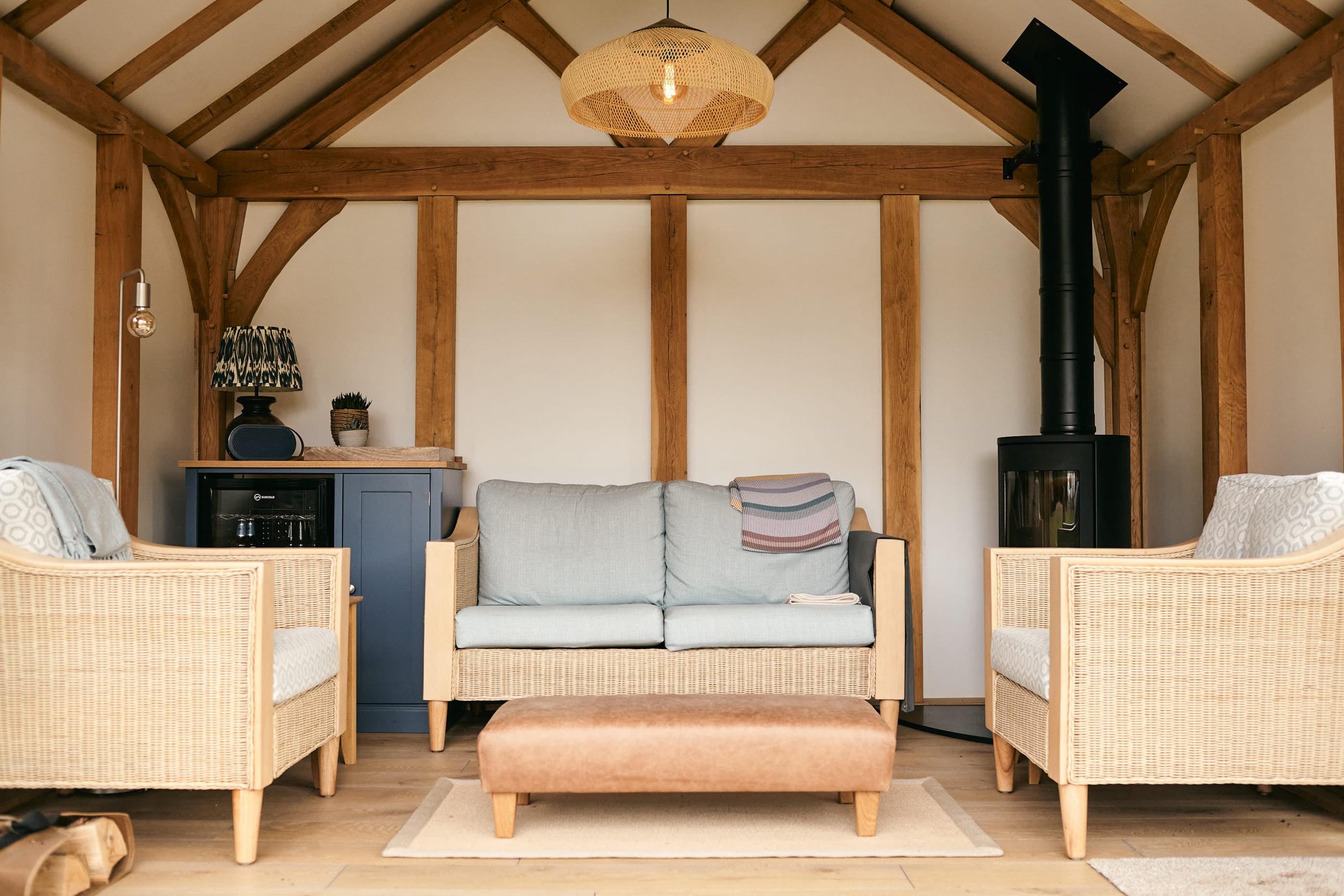 An oak framed garden studio, with a light gray sofa, two wicker armchairs, a brown ottoman, a blue cabinet with a lamp and a small plant, and a black wood stove.