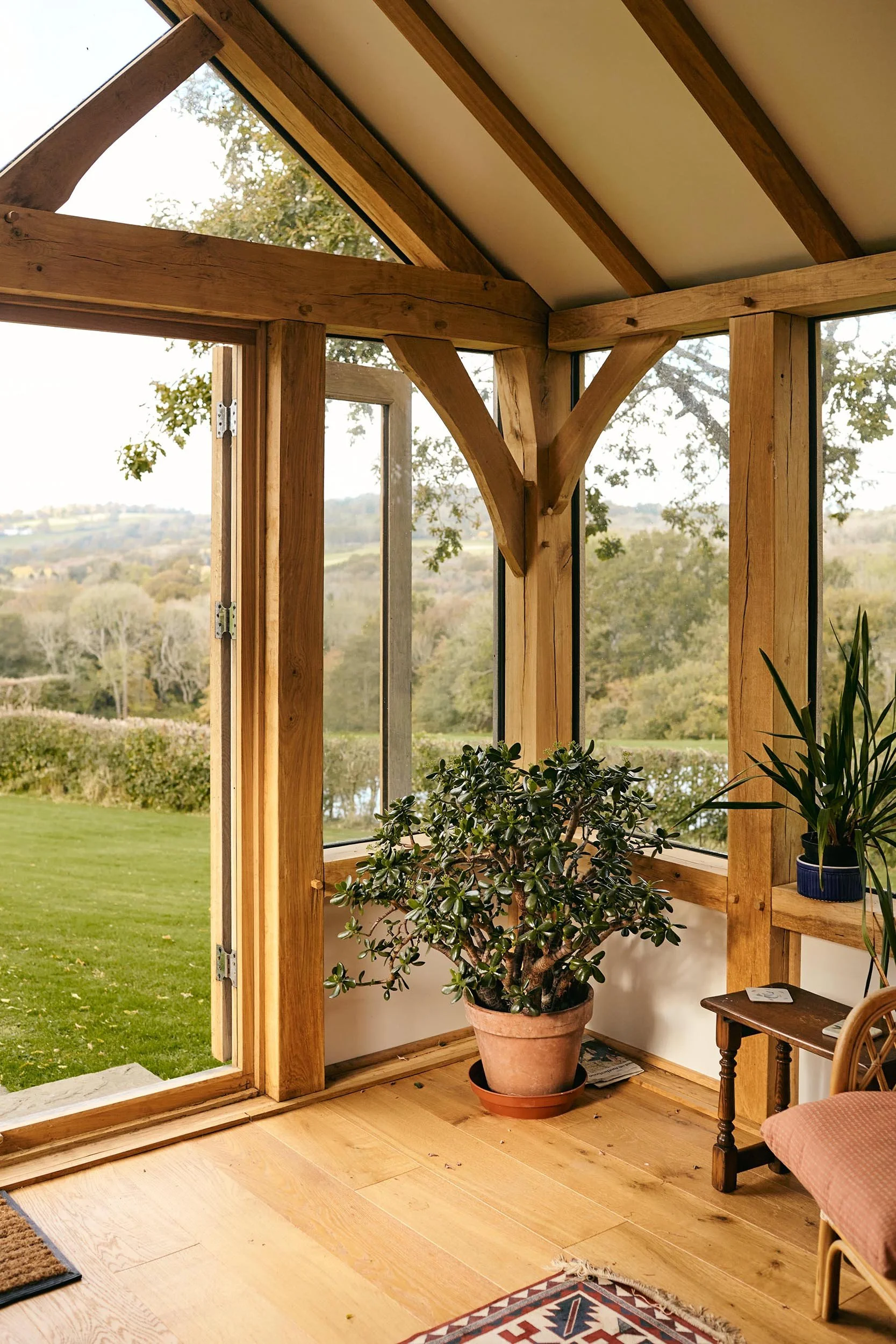 Interior view of an oak framed garden studio sunroom with large windows, wooden beams, a potted plant, a small side table, and part of a chair with a cushion, overlooking a grassy yard and trees outside.