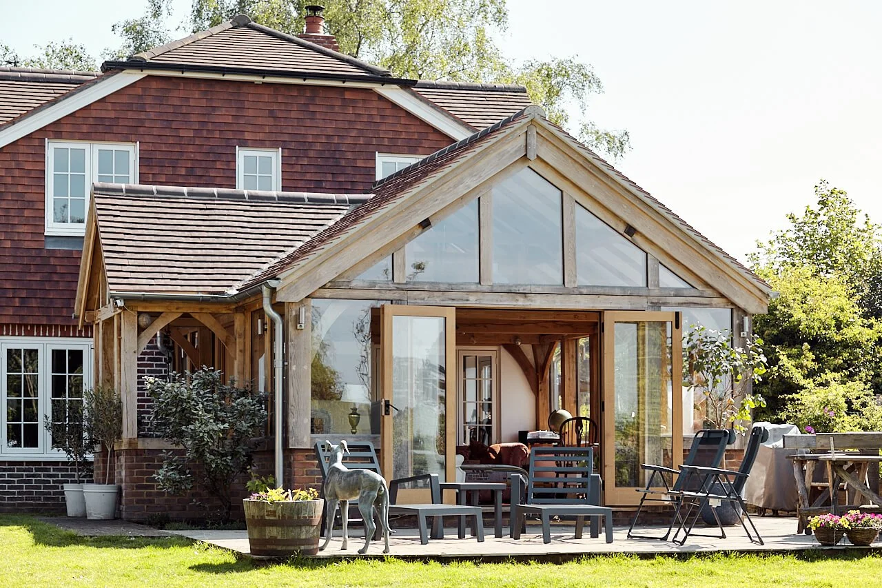 A house with a brick exterior and an oak framed extension and oak framed porch, outdoor furniture, and potted plants in a lush green backyard.
