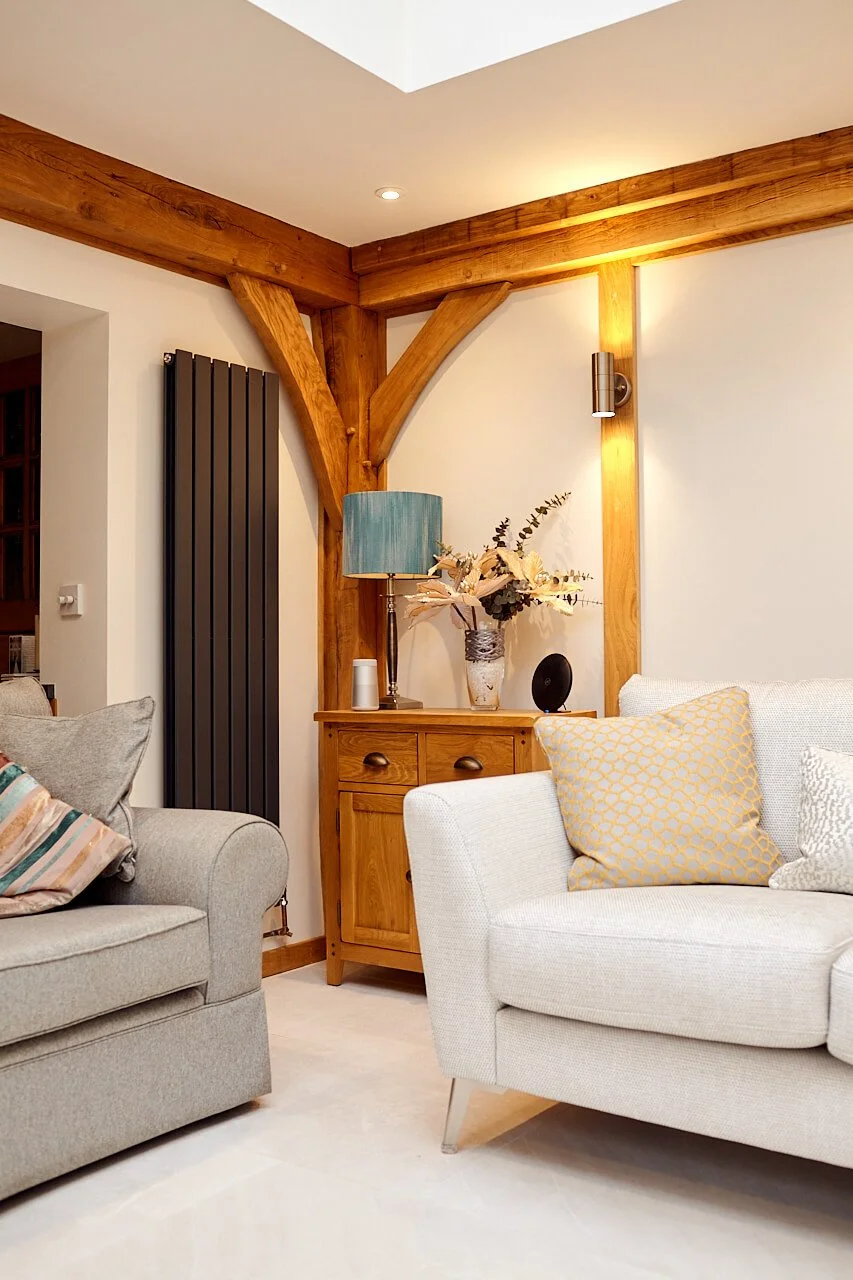 Living room in an oak framed orangery extension, with two beige sofas, wooden accents, a cabinet with a floral arrangement, a teal lampshade, and modern decor.
