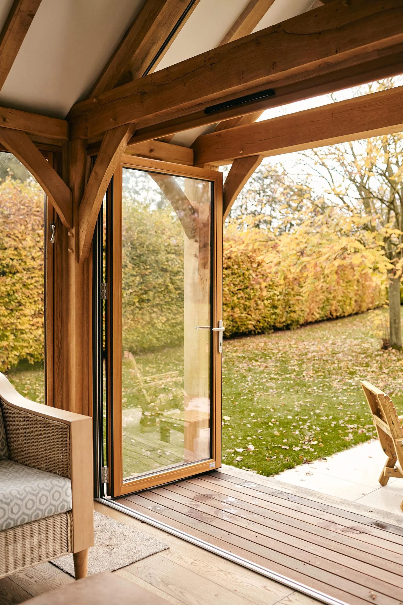 An open oak framed glazed bifold door and glass door leading to a back garden with fallen leaves and trees with autumn foliage.