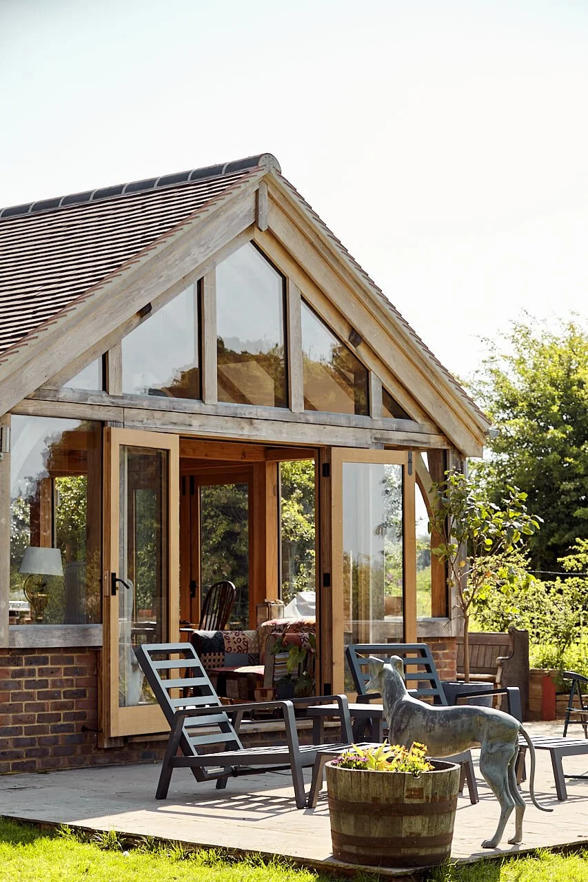 An oak framed house extension with large glass windows and doors, surrounded by outdoor seating, greenery, and a dog statue.