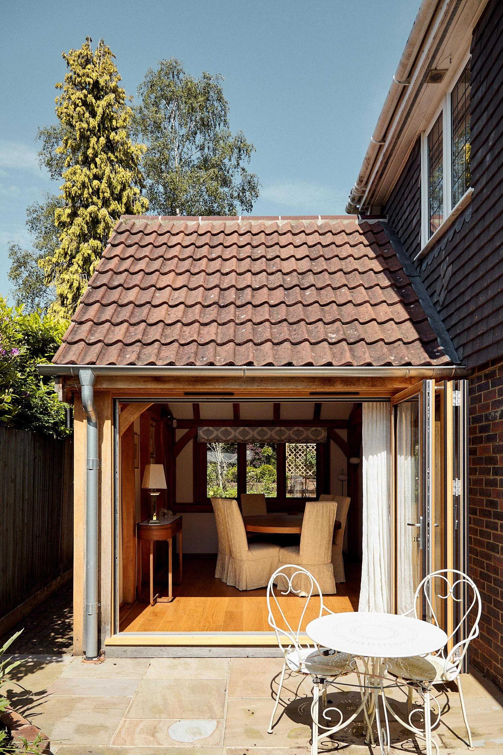 View of a back garden with a patio, metal table and chairs, and an oak framed sunroom with large windows and wooden interior, with trees in the background and a clear sky.