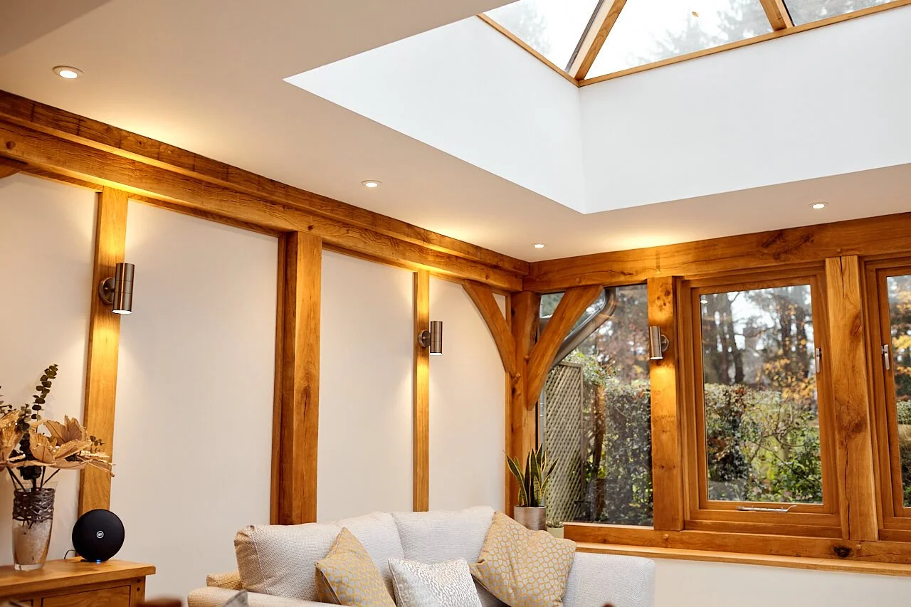 Living room in an oak framed orangery with wooden beams, a skylight, large windows showing trees outside, a white sofa with yellow and silver pillows, a potted plant, and a vase with dried flowers on a wooden side table.