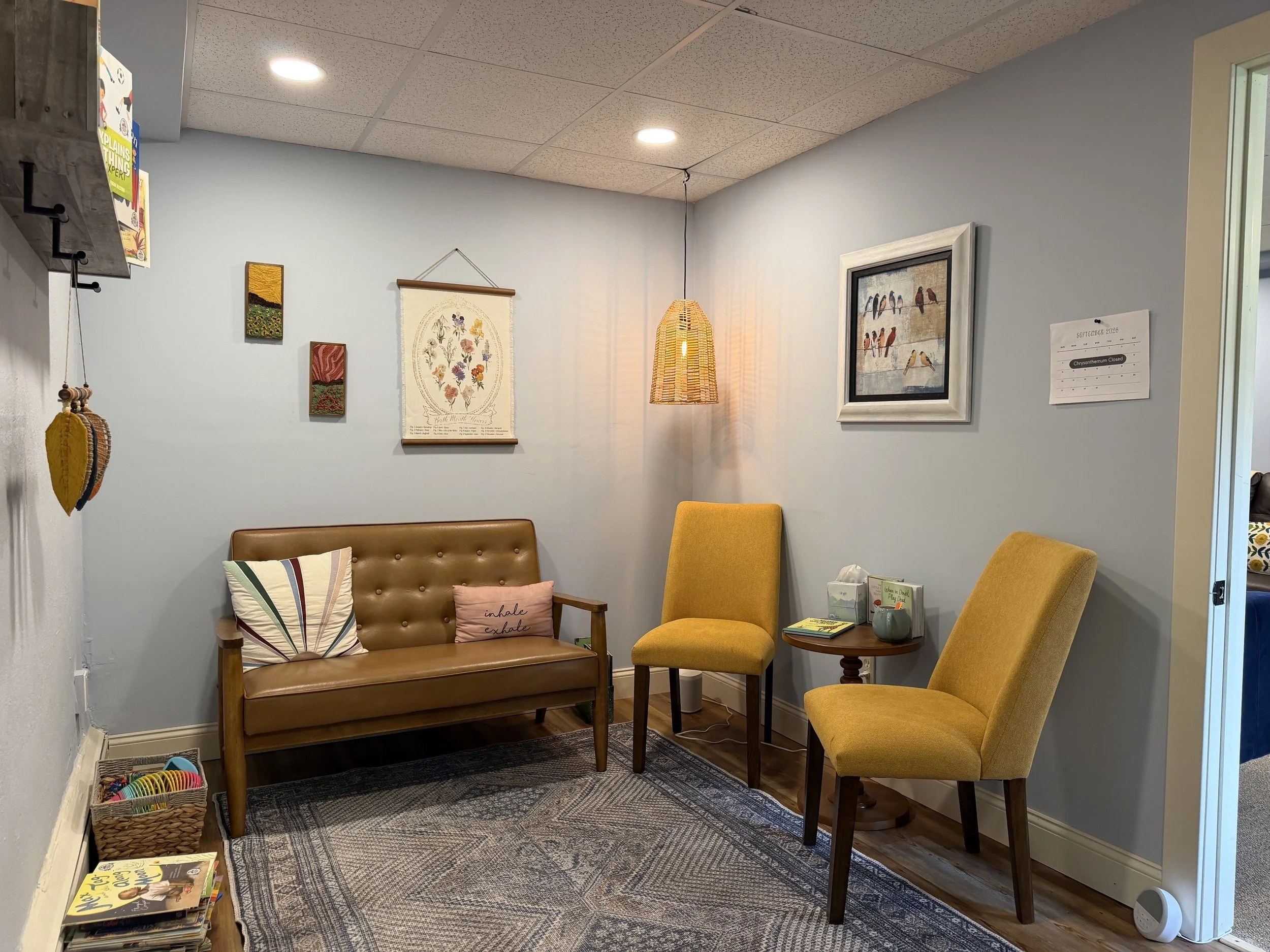 A cozy waiting area with a brown leather sofa, two mustard-colored armchairs, and a small wooden side table. The wall is decorated with framed artwork, including a bird picture and floral tapestry. A hanging wicker lamp provides warm lighting, and a small bookshelf with books and magazines is visible at the bottom left corner. The room has light blue walls and a patterned rug.