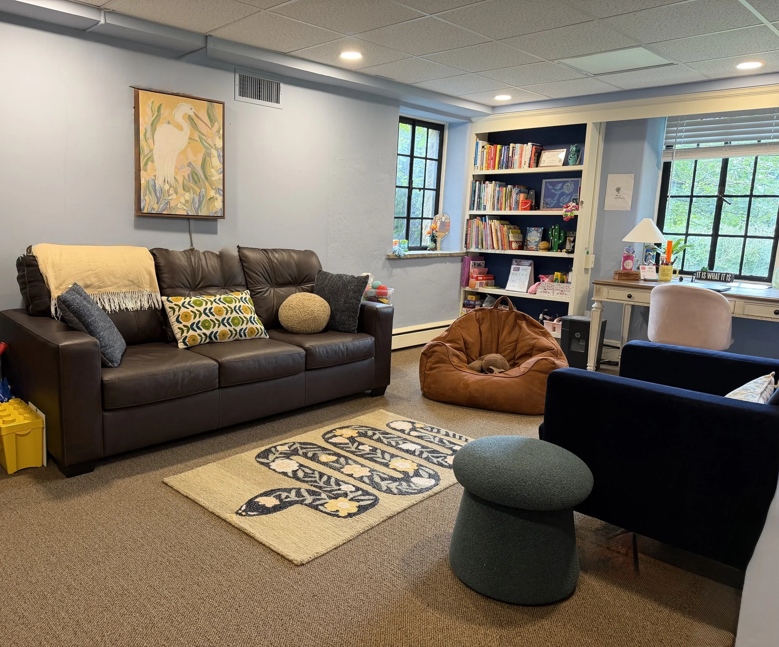 A cozy living room with a brown leather sofa decorated with patterned cushions, a beige knit throw, and a plush round cushion. There is a colorful area rug on the beige carpeted floor. A wine-colored stuffed animal is on the floor. The walls are painted light blue, and a painting of a white heron among green foliage hangs on one wall. To the right, there is a large window with black framing, letting in natural light. Next to the window, a tall bookshelf filled with books, framed pictures, and decorative items is visible. A small table below the window holds various items, including a lamp, a plant, and some stationery.