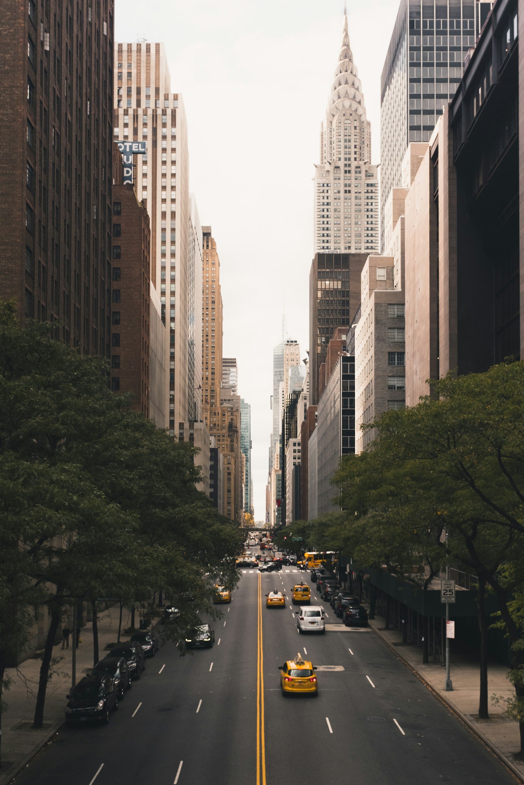 A city street view with tall buildings on both sides, yellow taxis and cars on the road, and trees lining the sidewalks, with the Chrysler Building visible in the distance in New York City.