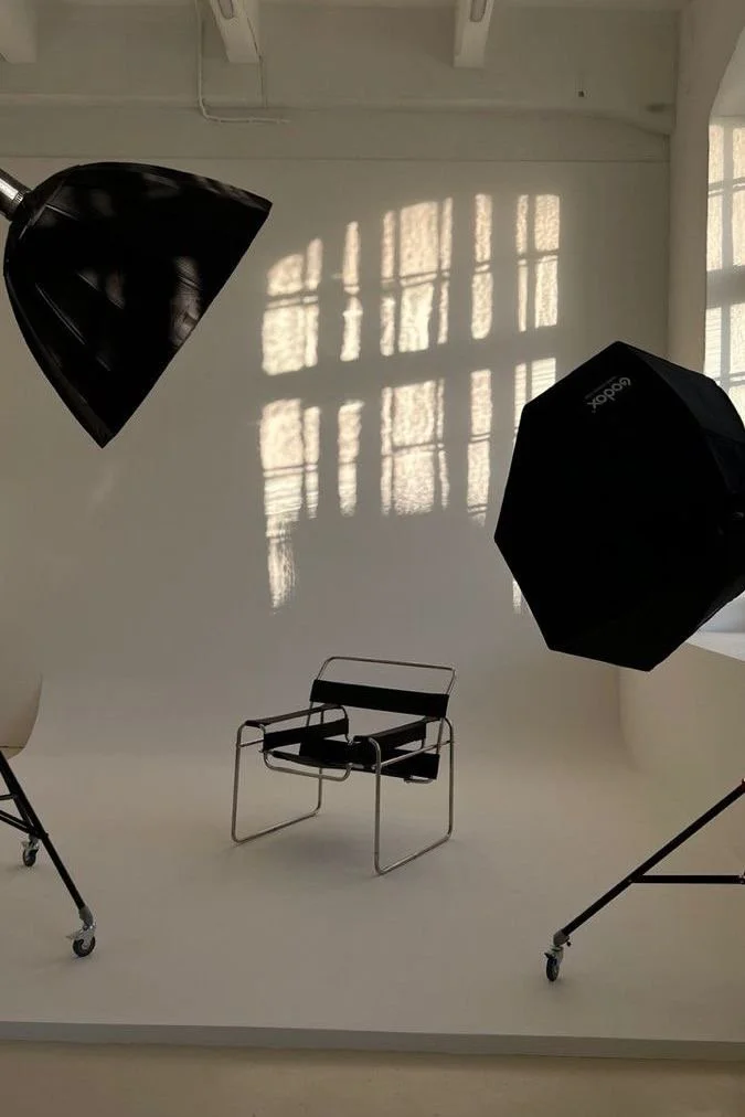 A photo of a photography studio with two large softbox lights and a black chair on a white backdrop, with window sunlight creating shadows on the wall.