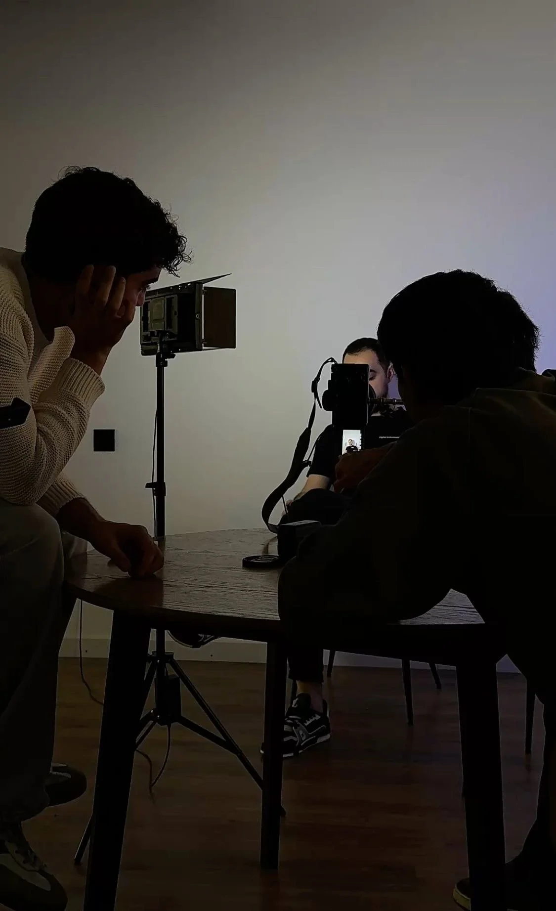 Three people in a room with a table and camera equipment; two are standing and one is sitting, involved in a filming or photo shoot setup.