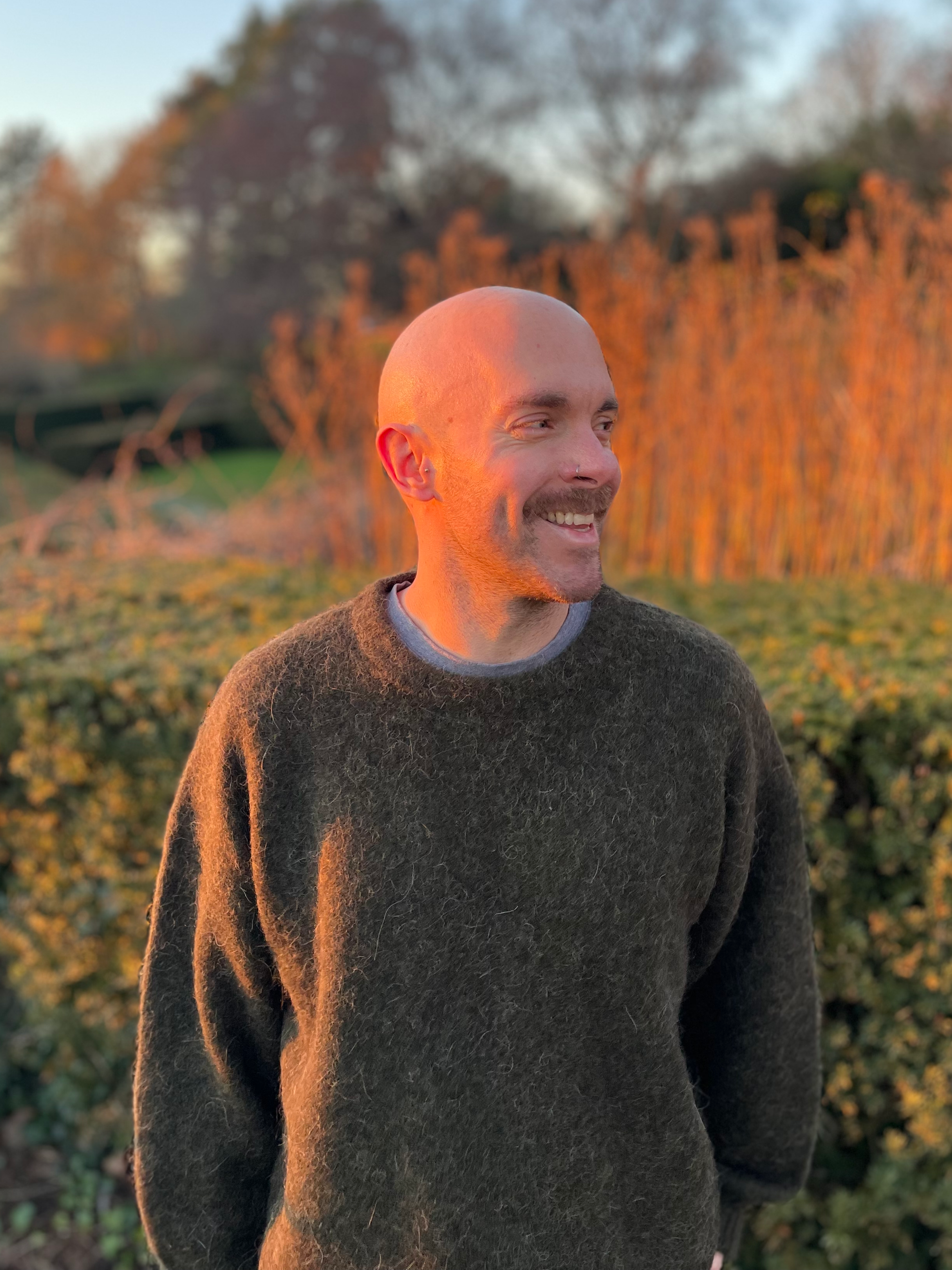 A smiling man with a shaved head and nose piercing, wearing a dark green jumper, standing outdoors during sunset with autumn trees and greenery in the background.