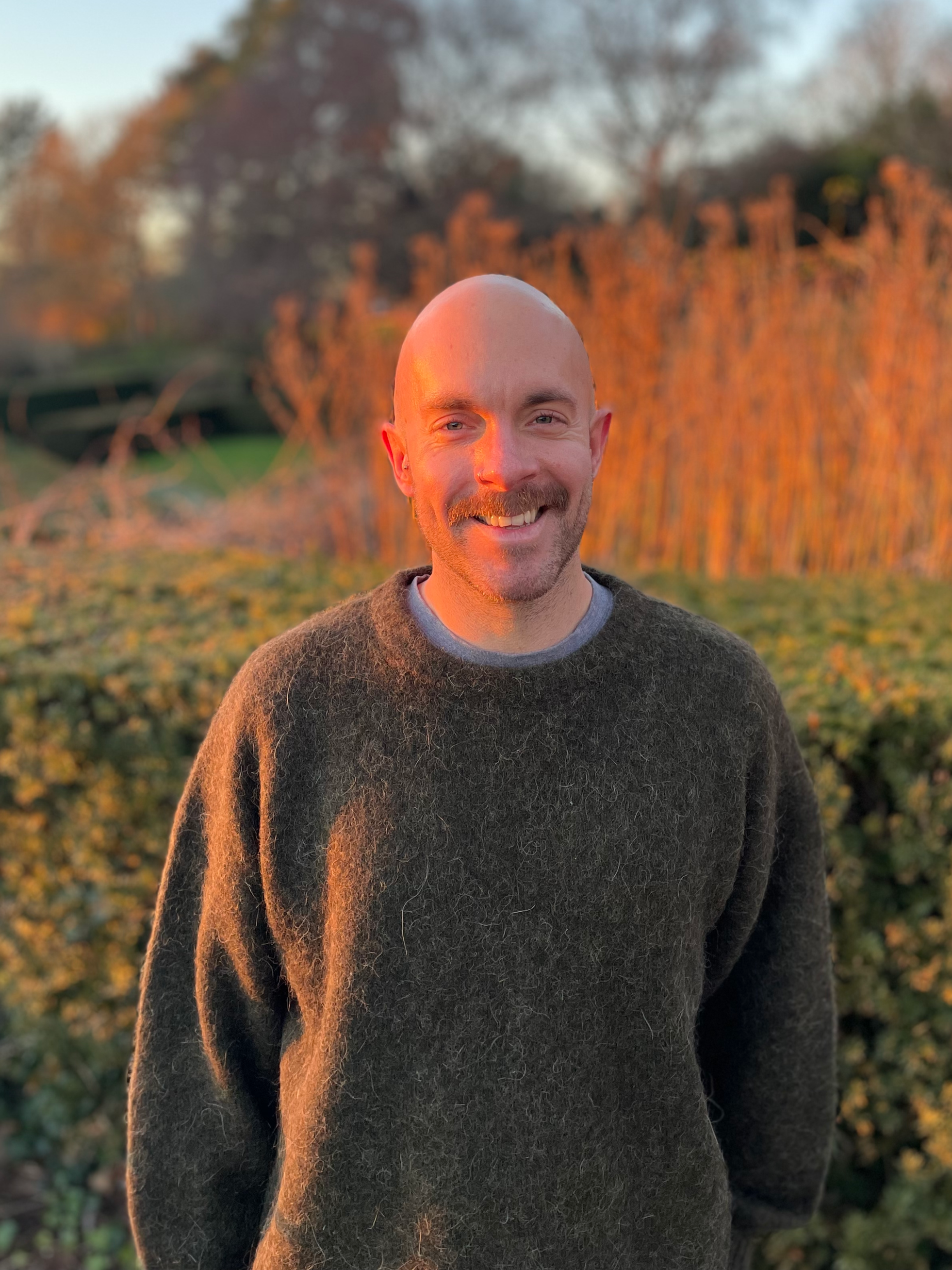 A man with a shaved head and a moustache smiling outdoors during sunset, standing in front of bushes and trees.