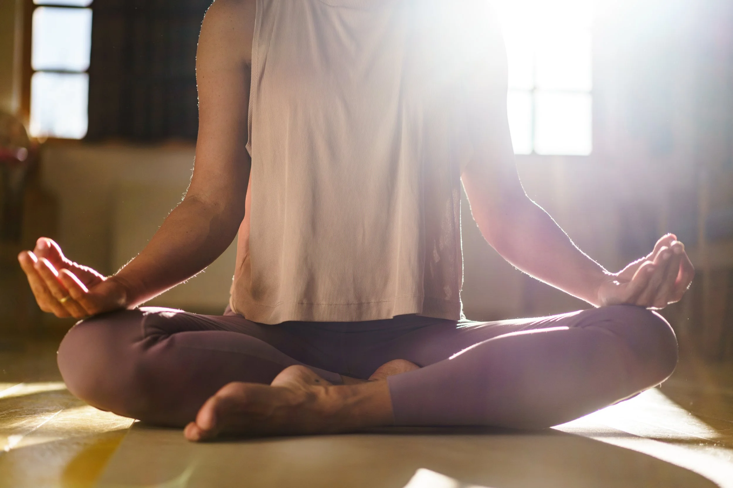Person meditating in a cross-legged sitting position on a yoga mat, with sunlight streaming through windows in the background.
