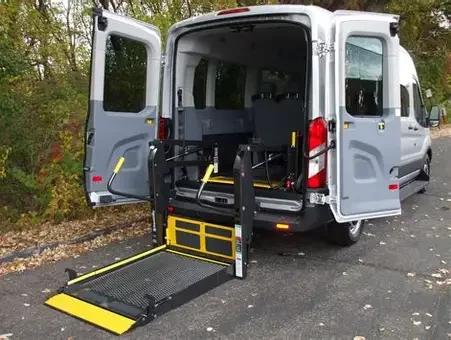 A silver van with its rear doors open, equipped with a wheelchair lift and ramp for accessibility parked on a paved surface surrounded by trees.