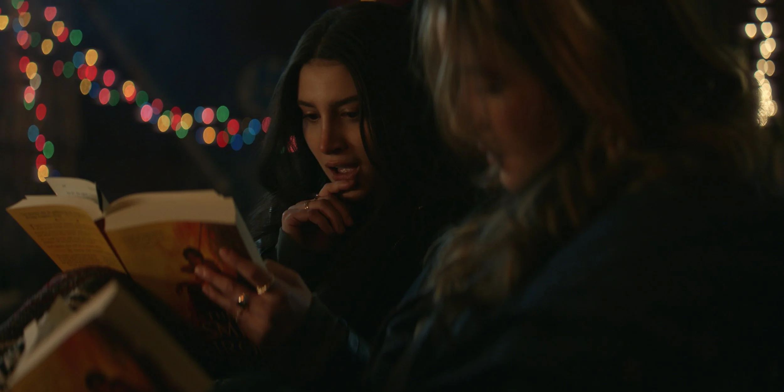 Two women reading books at a nighttime gathering with colorful string lights in the background.