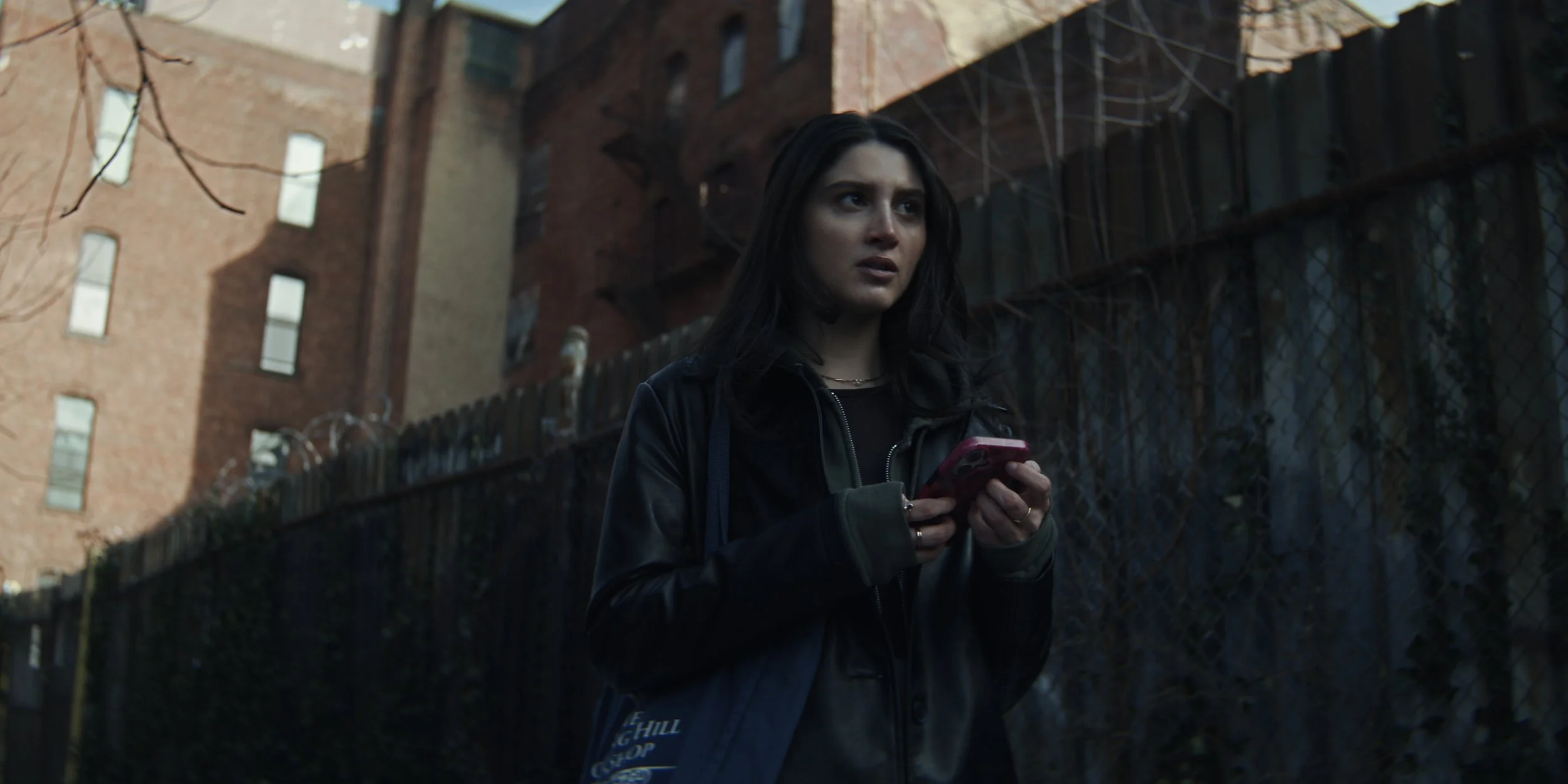 A young woman with black hair, wearing a black jacket, standing outdoors near a metal fence and brick apartment buildings, holding a smartphone and looking worried or focused.