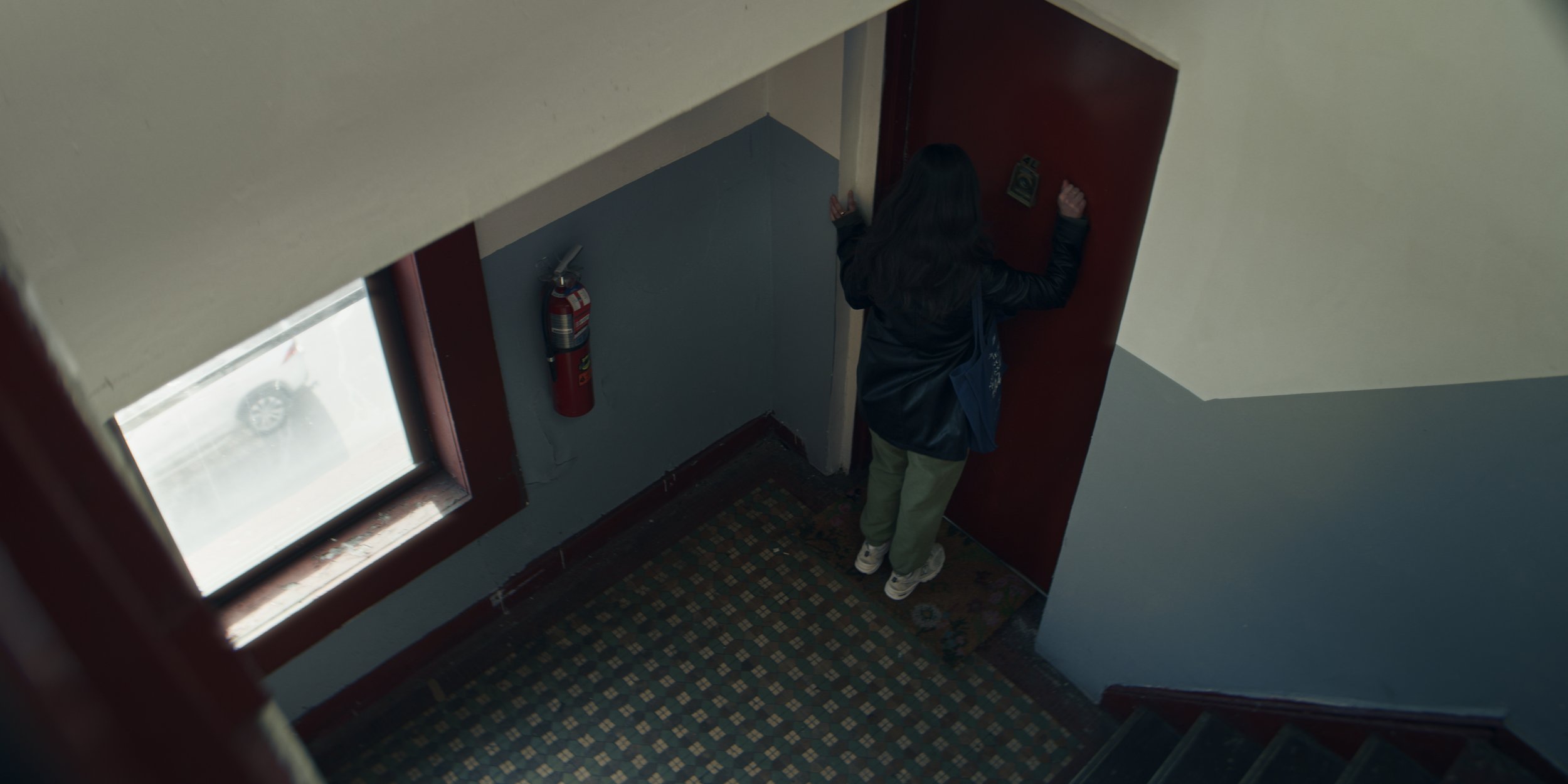 A woman with dark hair, wearing a black jacket, green pants, and white shoes, stands at a red door in a stairwell, looking away from the camera. There is a fireplace on the wall and a window with snow outside. A fire extinguisher is mounted on the wa