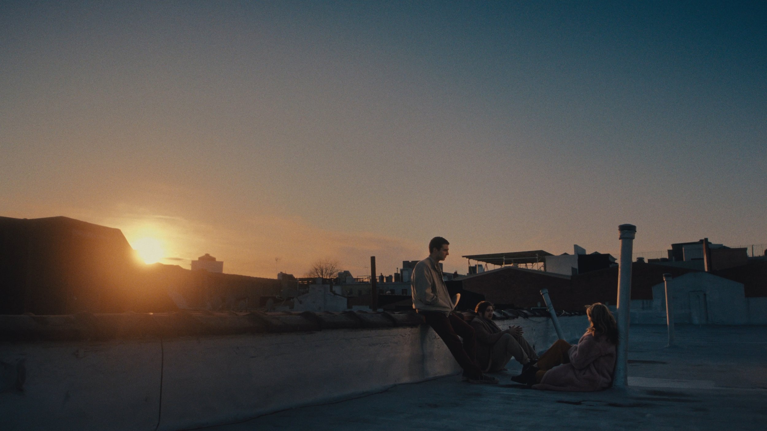 Three people sitting and talking on a rooftop at sunset, with the city skyline and a clear sky in the background.