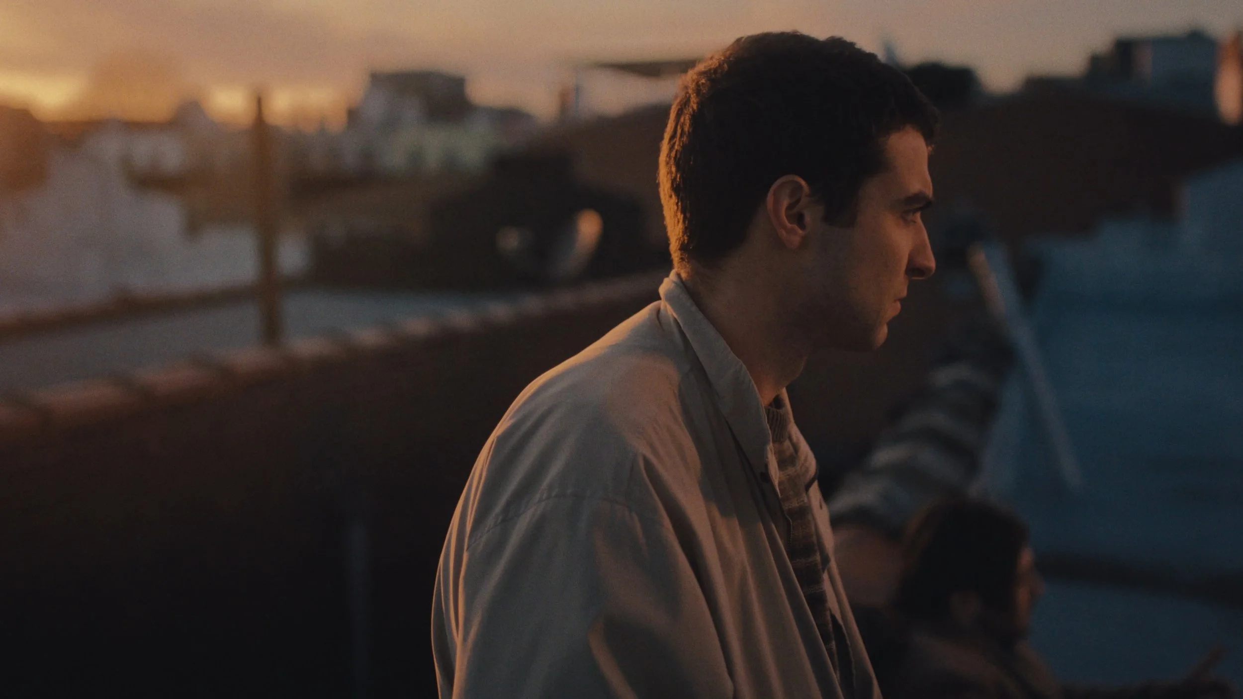 A man in profile sits outdoors during sunset, on a roof with  buildings in the background.