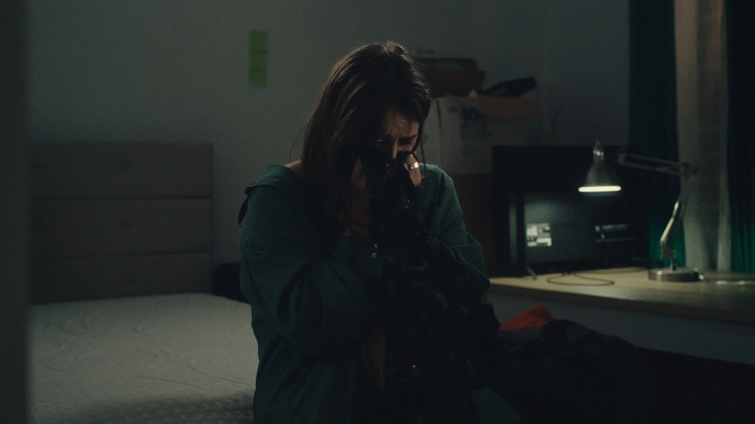 A woman sitting on a bed, holding her face with her hands, appearing distressed in a dimly lit room.