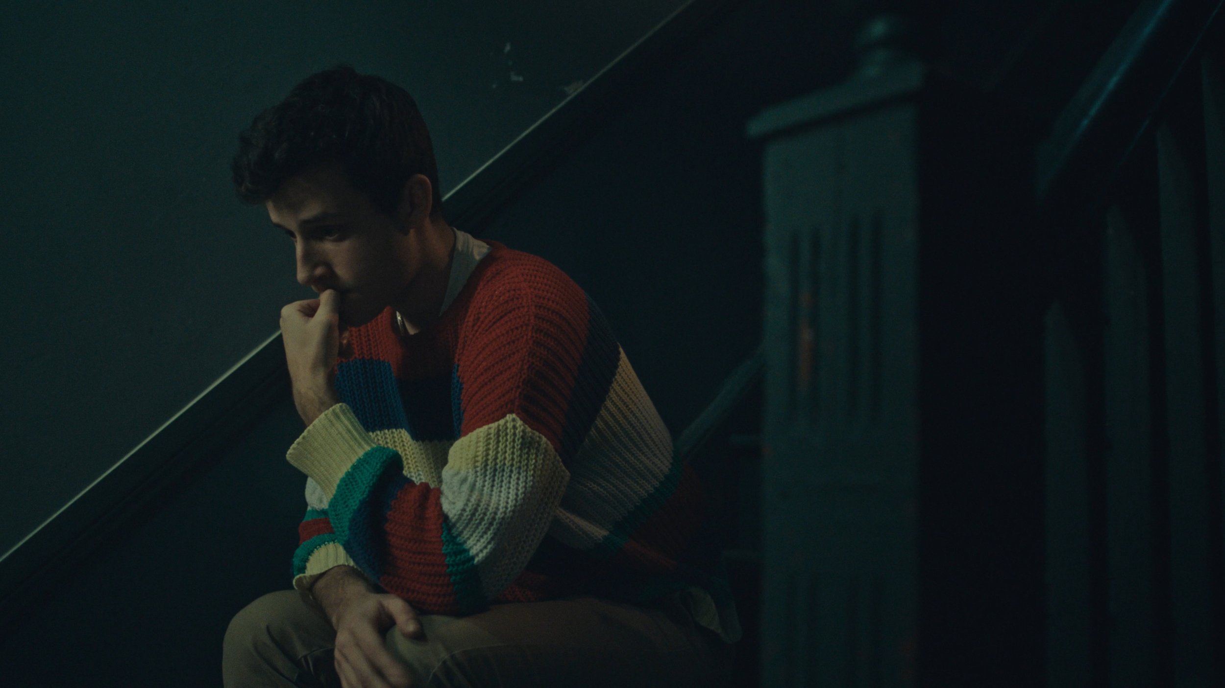 A young man sitting on stairs in dim lighting, wearing a colorful striped sweater, with a contemplative or worried expression, resting his chin on his hand.