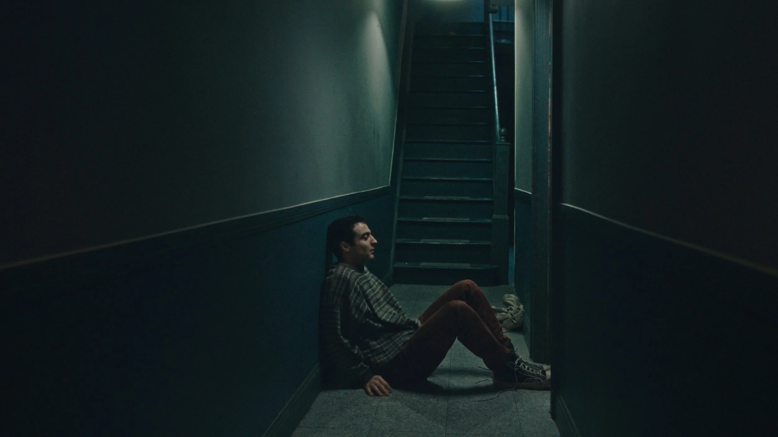 A person sitting on the floor of a dimly lit hallway with dark walls and a staircase in the background, looking to the left.